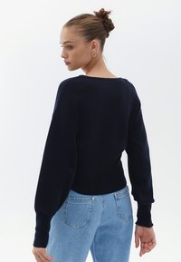 Marinblå stickad tröja med rundad halsringning och långa, något puffiga ärmar. Kombinerad med ljusblå jeans som visar en avslappnad passform och mjuk textur.