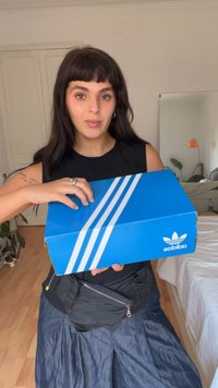 Kék Adidas cipősdoboz három fehér csíkkal, egy személy tartja. A doboz oldalán logó található. Fa padló és világos háttér.