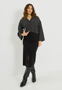 Blouse noire à pois avec manches larges, associée à une jupe noire cintrée avec une fente avant. Le modèle tient une petite pochette noire.