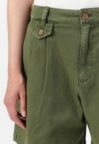 Calções de algodão verde-azeitona com cintura alta, apresentando bolsos laterais e um detalhe de aba com botão na parte frontal. Textura suave e corte ajustado.