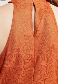 Närbild av en persons övre rygg som bär en ärmlös, strukturerad orange klädesplagg med en öppning i nacke mitt bak.