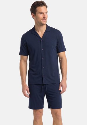 Mand iført navy blå kortærmet skjorte med knapper og matchende shorts, stående og kiggende let til siden mod en ensfarvet baggrund.