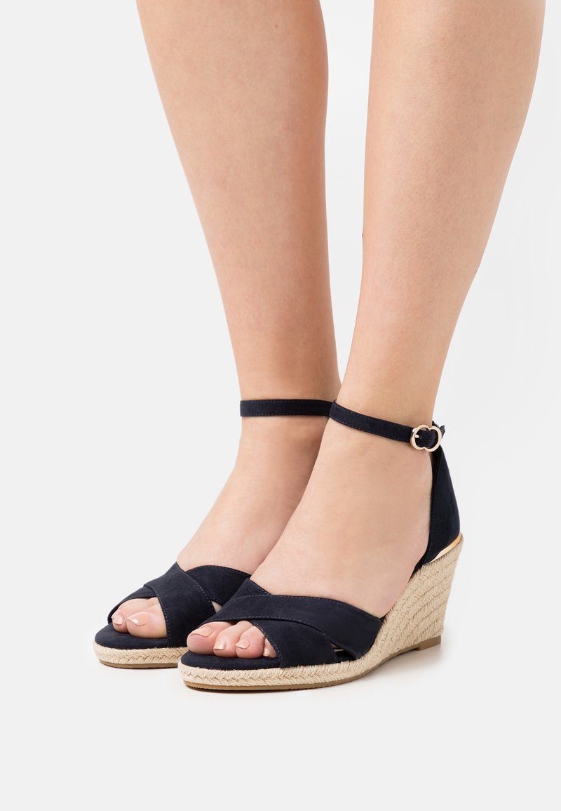 Ben som bär marinblå kilklackar med ankelsnören och vävda repklackar mot en vit bakgrund.