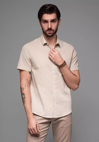 Lichtbeige korte mouw button-up shirt, zachte textuur, puntige kraag, enkele zak aan de linkerkant, gedragen met lichtgekleurde broek.