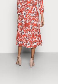 Robe midi rouge à fleurs avec des manches longues, présentant une taille froncée et des motifs floraux blancs. Assortie à des escarpins nude à bride cheville.