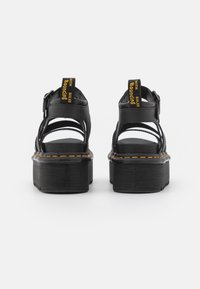 Des sandales noires à plateforme présentent un design ouvert avec plusieurs sangles, une semelle en caoutchouc texturée et une étiquette jaune proéminente sur le talon.