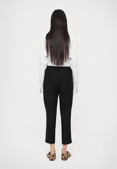 Pantalon noir ajusté avec poches latérales, associé à une chemise blanche à manches longues. Les chaussures présentent un motif léopard.