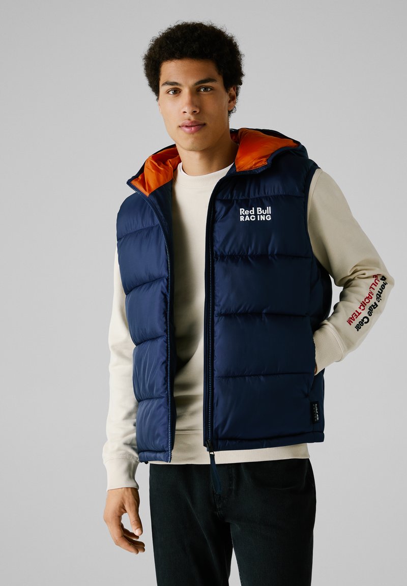 Chaleco azul marino acolchado con capucha naranja, cremallera frontal y logo de Red Bull Racing. Lleva puesto sobre un suéter de color claro con detalles en las mangas.