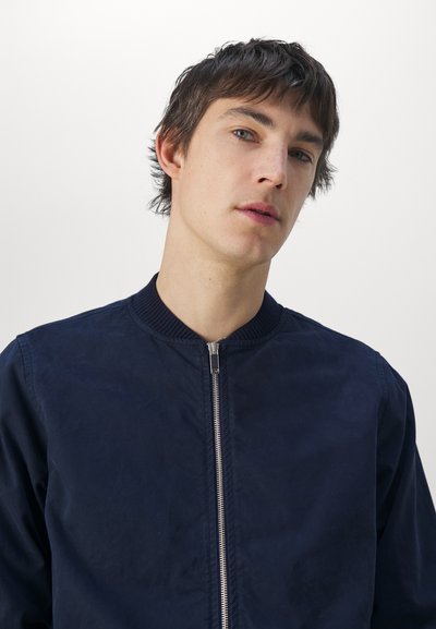 Chaqueta de bomber navy con acabado mate, que presenta un cuello acanalado y una cremallera frontal. Diseño limpio sin patrones ni adornos.