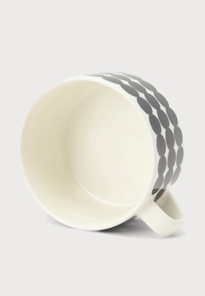 Бяла керамична чаша, легнала настрани, с дръжка и три реда големи черни точки около външната страна.