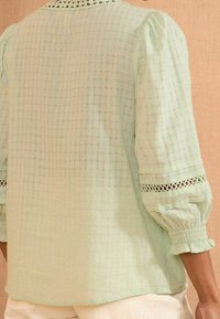 Jasnozielona, teksturowana bluzka z bufiastymi, trzyćwiartkowymi rękawami i koronkowym wykończeniem przy elastycznych mankietach, noszona z białymi spodniami.
