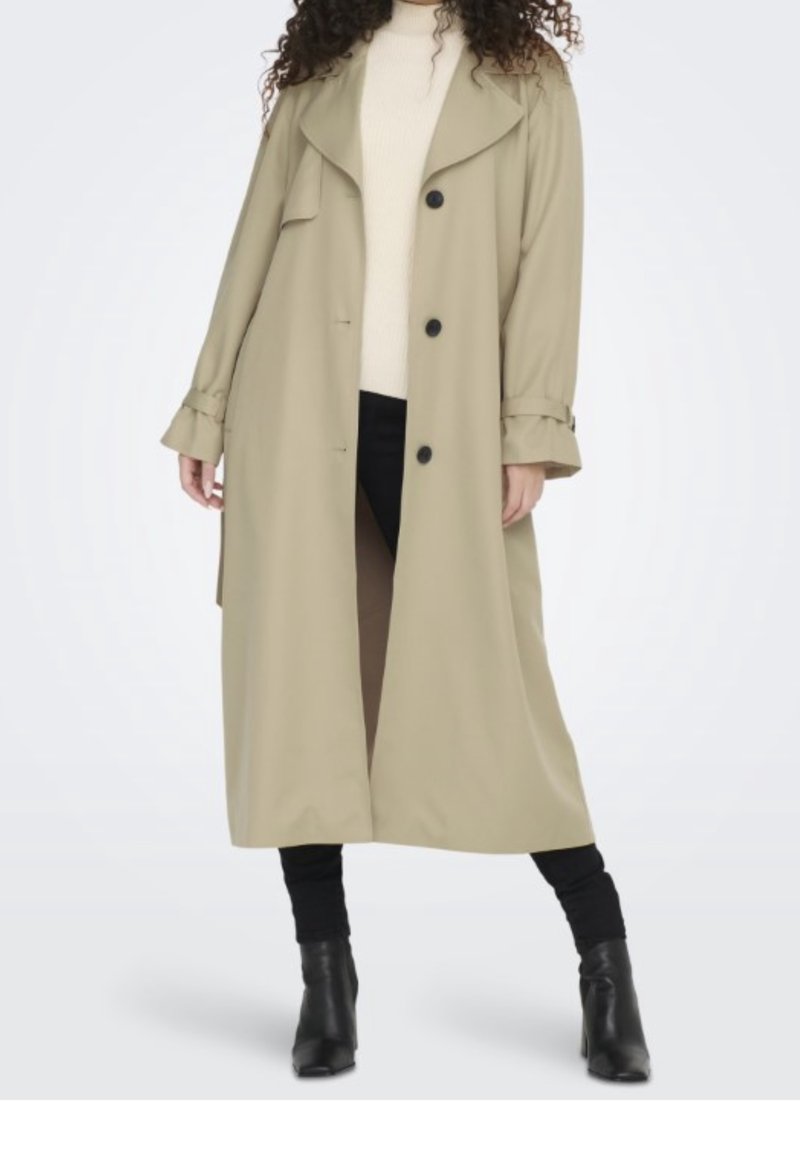 Personne portant un long trench-coat beige sur un haut blanc et un pantalon noir avec des bottines noires, debout sur un fond blanc.