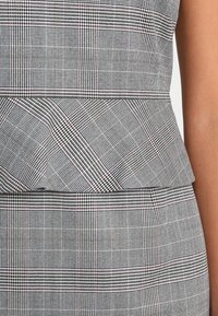Gros plan d'un vêtement ajusté en tissu à carreaux noir et blanc avec de subtiles lignes rouges, montrant un design en couches autour de la taille et des hanches.