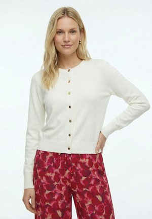Vrouw met blond haar die een witte, geknoopte cardigan en rode, bloemenbedrukte broek draagt, staat met één hand op haar heup.