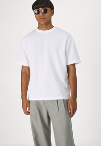 Weißes Kurzarm-T-Shirt, lässige Passform, aus Baumwolle, kombiniert mit grauen geplätteten Hosen; das Model trägt runde Sonnenbrillen.