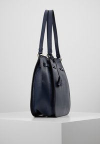 Laukkua tummansinisestä nahasta, jossa on mukulakivimäinen rakenne, leveä runko, kaksinkertaiset olkahihnat ja metalliset yksityiskohdat. Sivuprofiili, minimalismi.