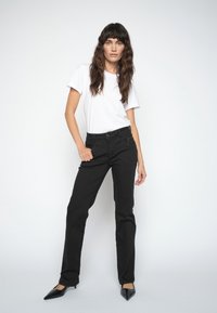 Frau mit langen braunen Haaren, die ein weißes T-Shirt, schwarze Hose und schwarze spitze High Heels trägt, steht vor einem einfarbig weißen Hintergrund.