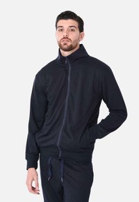 Giacca nera con zip e colletto alto, texture morbida e maniche lunghe. Presenta tasche laterali e una vestibilità aderente. Abbinata a pantaloni coordinati.