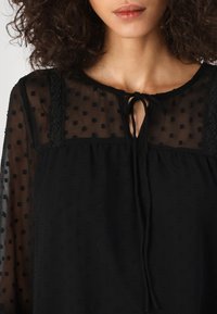 Blusa nera in tessuto trasparente a pois, scollatura rotonda, delicato dettaglio con fiocco e accenti in pizzo testurizzato sulle spalle. Materiale morbido e leggero.
