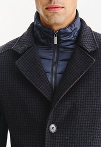 Granatowa kurtka puchowa z kołnierzem, uszyta z ciemnego materiału w kratę, z zamkiem błyskawicznym z przodu, dużymi klapami i zapięciem na guziki.