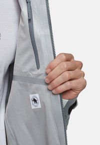 Lys grå jakke indvendig med lynlås lomme, synlig etiket og tekstil med struktur; har kontrasterende grå kant og en glat for.