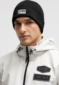 Black ribbed knit beanie featuring a stitched label with "CAMP DAVID." Worn with a light-colored jacket with multiple patches and zippers.