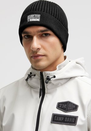 Black ribbed knit beanie featuring a stitched label with "CAMP DAVID." Worn with a light-colored jacket with multiple patches and zippers.