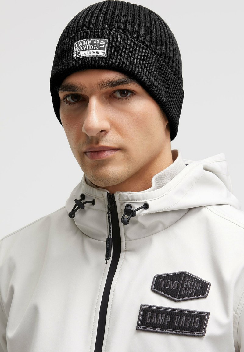 Black ribbed knit beanie featuring a stitched label with "CAMP DAVID." Worn with a light-colored jacket with multiple patches and zippers.