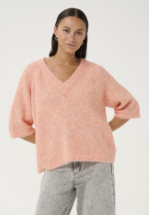 Lichtroze, oversized V-hals gebreide trui met korte mouwen, zachte textuur en een iets cropped afwerking. Combinatie met grijze jeans.