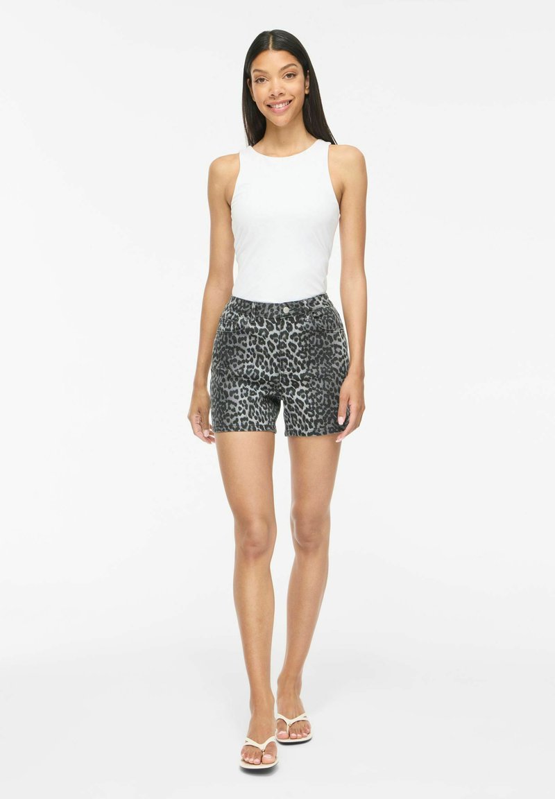 Femme debout, portant un haut blanc sans manches, un short gris à motif léopard et des sandales blanches, souriante sur un fond blanc uni.