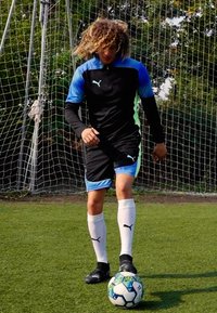Schwarz-blaues Sporttrikot mit kurzen Ärmeln, kombiniert mit schwarzen Shorts und weißen Socken, stehend auf Gras, neben einem Fußball.