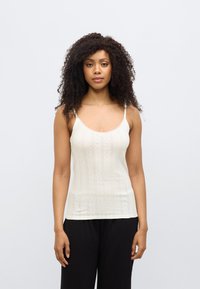 Débardeur crème à fines bretelles, avec un motif texturé et une bordure en dentelle le long de l'encolure. Porté avec un pantalon noir.