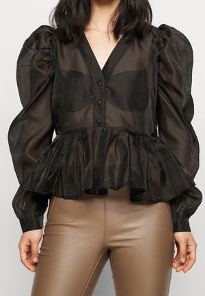 Frau trägt eine transparente schwarze Bluse mit Puffärmeln, Knöpfen und Peplum-Saum, kombiniert mit hochgeschnittenen beigen Lederhosen.