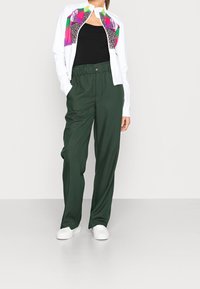 Witte jas met kleurrijke bloemenpatronen, zwarte top, groene wijde broek en witte sneakers. Heeft een relaxte pasvorm en elastische tailleband.