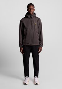 Grå, letvægtsjakke med hætte, der har elastiske manchetter og et gult logo på brystet, kombineret med sorte bukser og hvide sneakers.