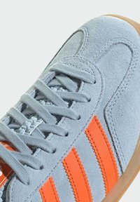Light blue suede sneaker with orange zigzag stripes, textured surface, and traditional lacing. Rubber outsole visible at the bottom.