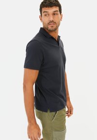 Marineblaues kurzärmeliges Polohemd mit einem Kragen, geripptem Muster und einer Brusttasche. Getragen mit hellgrünen Cargo-Shorts.
