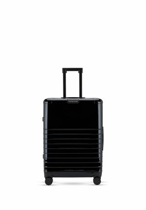 Valise rigide noire avec des rainures horizontales, quatre roues et une poignée télescopique étendue sur fond blanc.