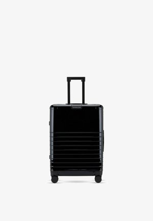 Valise rigide noire avec des rainures horizontales, quatre roues et une poignée télescopique étendue sur fond blanc.