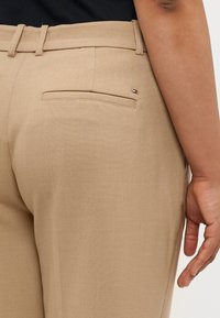 Lys beige bukser med en glat tekstur, en baglomme med en vandret åbning og en lille logo accent nær taljen.