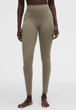 Mørkhudet person iført højtaljede, olivengrønne tætsiddende leggings, stående barfodet på en ensartet lys grå baggrund.