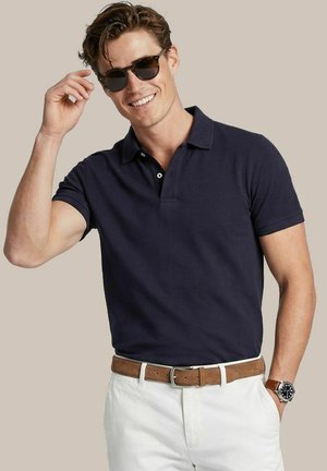 Glunderende man die zwarte zonnebril draagt, een donkerblauw poloshirt, witte broek met bruine riem en polshorloge, en met één hand zijn zonnebril verstelt.