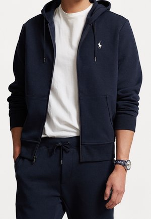 Mann trägt eine marineblaue Zip-up-Kapuzenjacke mit Kordeln, ein weißes T-Shirt, passende marineblaue Hose und eine Uhr mit marineblauem Armband am linken Handgelenk.