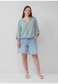 Hellbluse mit V-Ausschnitt und hochgerollten Ärmeln, kombiniert mit zerrissenen Jeansshorts. Das Model trägt weiße Riemchensandalen.