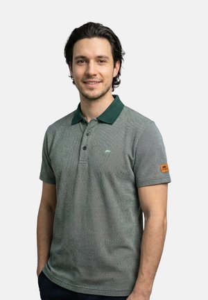 Mann mit kurzen dunklen Haaren, der ein graues Poloshirt mit dunkelgrünem Kragen trägt, lächelt mit den Händen in den Taschen vor einem einfarbigen Hintergrund.