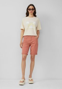 Creme graphic t-shirt met palmboom- en zonontwerp, gecombineerd met koraalkleurige opgerolde shorts en platformsandalen met brede banden.