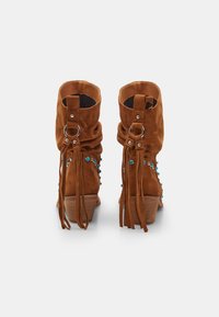 Botas de ante marrón hasta el tobillo con un diseño holgado, detalles de flecos, herrajes metálicos y acentos de cuentas azules en los lados exteriores. Tacón bajo en bloque.