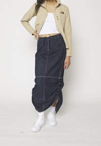 Denimkjol med dragsko i midjan, blå färg, med kontrasterande sömmar och en sidofrak. Matchad med en beige jacka och vita sneakers.