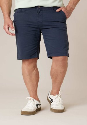 Homme portant un short bleu marine et des baskets blanches, debout avec une main dans la poche devant un fond uni.