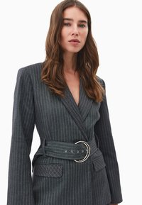 Blazer gris a rayas diplomáticas a medida con solapas anchas, mangas largas, una cintura con cinturón con una hebilla circular plateada y bolsillos frontales.